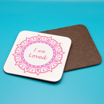 White positive affirmation coaster with pink and red mandala design. Daily affirmation reads I am Loved. Coaster underneath shows wood backing