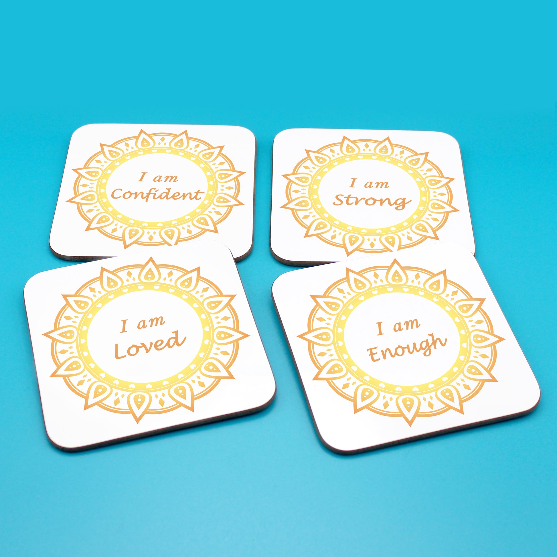 Wellbeing gift. Four square wooden backed personal affirmations coaster set. Mandala design with daily affirmation wording inside the mandala.  Each Affirmations coaster individually reads I am Loved, I am Enough, I am Confident, I am Strong (all mandalas are yellow and orange in this set)