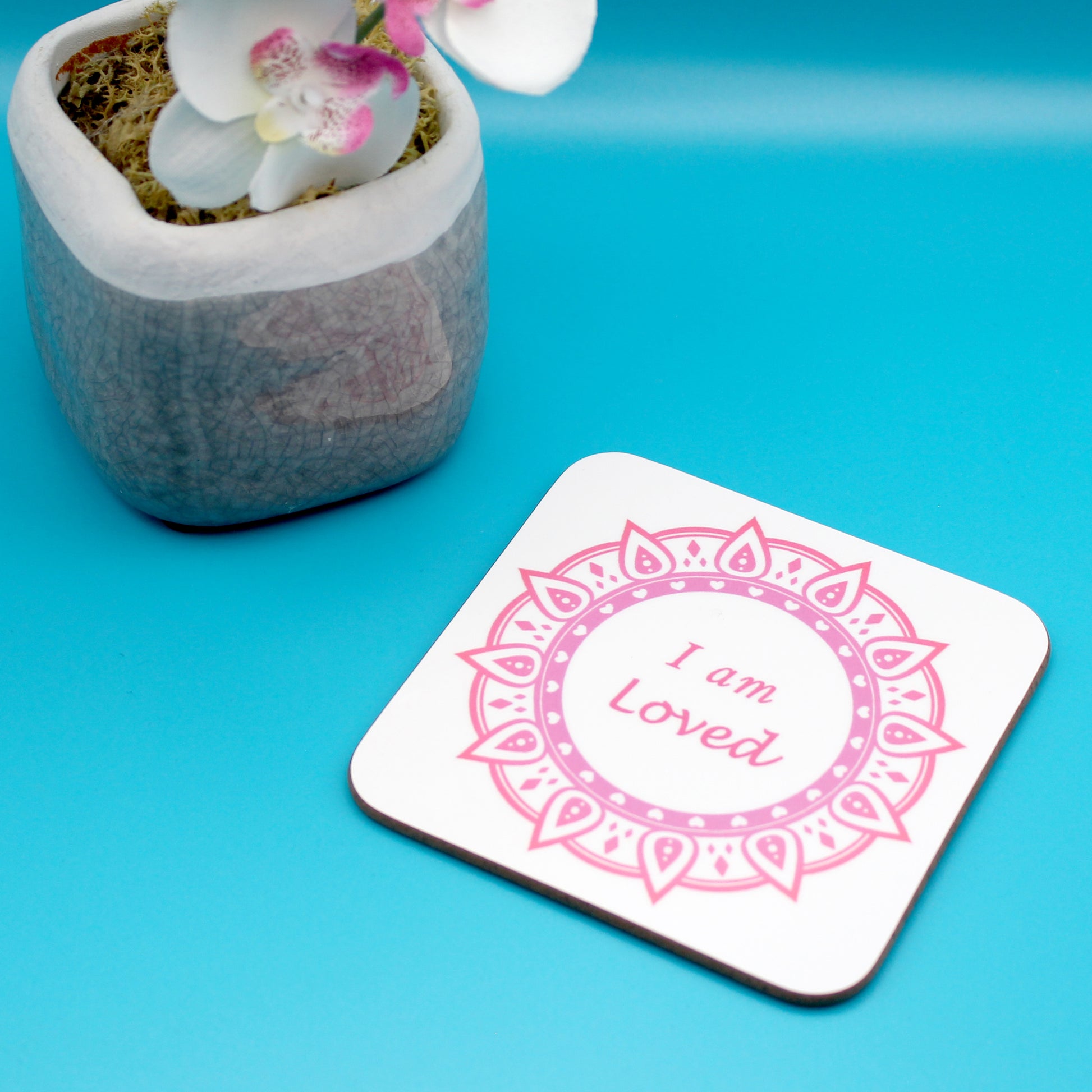 White positive affirmation coaster with pink and red mandala design. Daily affirmation reads I am Loved. Coaster underneath shows wood backing. Blue background with potted pink orchid to left corner.