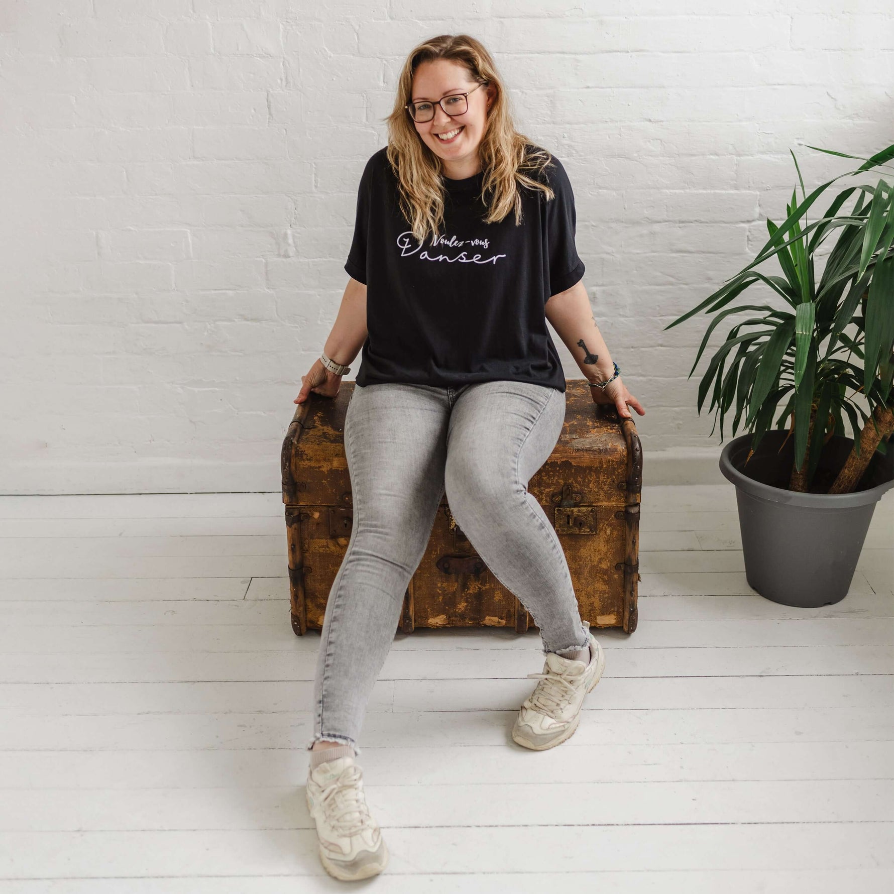 Woman wearing a black T-shirt with loose sleeves with roll cuffs worn with grey skinny jeans. The ladies tshirt has the words Voulez Vous Danser in white  She is sitting on a wooden weathered chest and is wearing white trainers