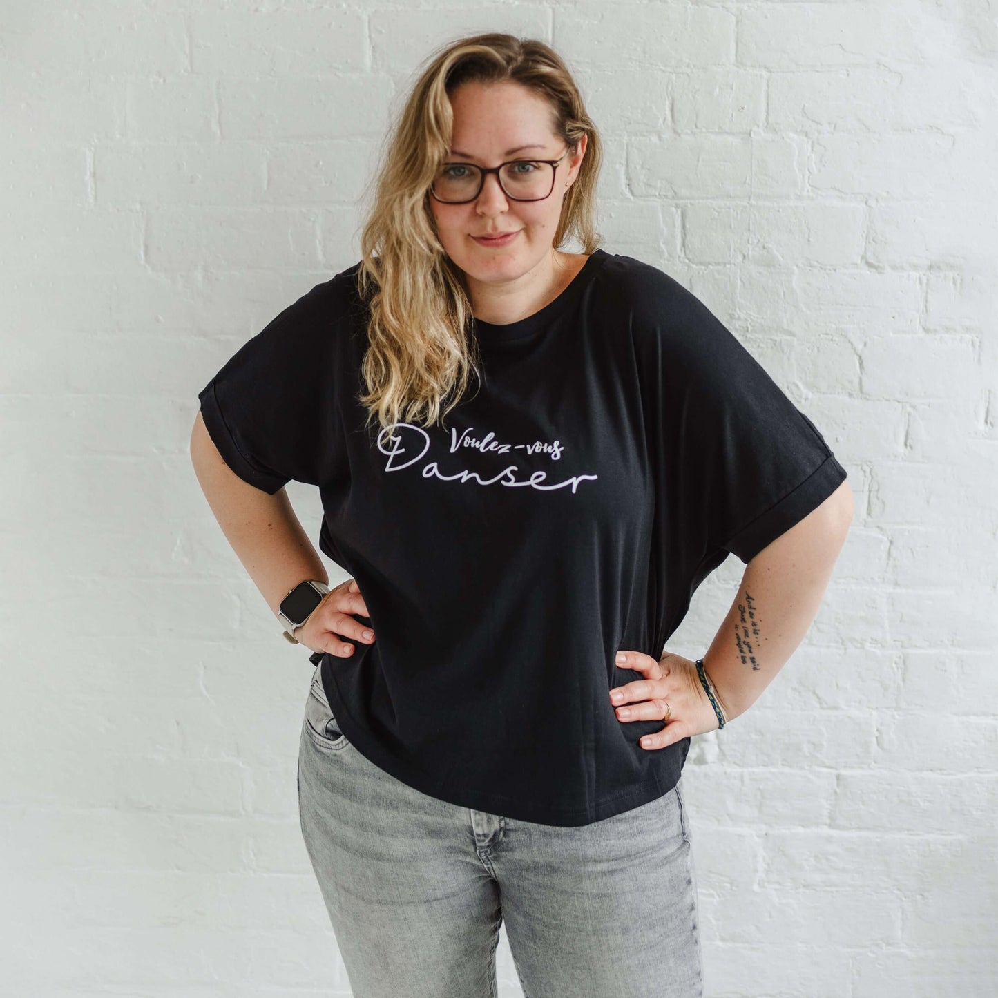 Woman wearing a black T-shirt with loose sleeves with roll cuffs worn with grey skinny jeans. The ladies tshirt has the words Voulez Vous Danser in white  Edit alt text