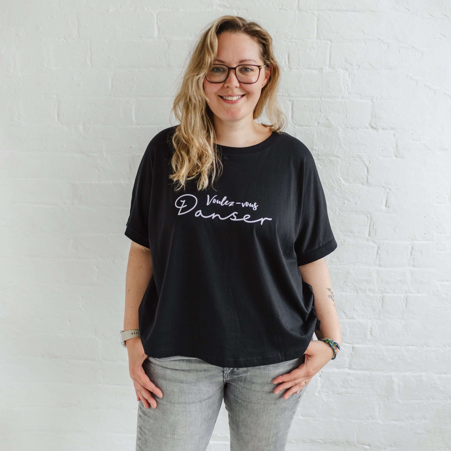 Woman wearing a black T-shirt with loose sleeves with roll cuffs worn with grey skinny jeans. The ladies tshirt has the words Voulez Vous Danser in white 