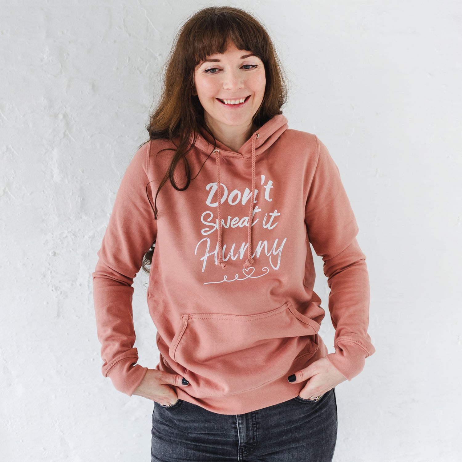 Woman wearing dusky pink fitted fashion womens long sleeve hoodie. Hoodie top for workout / casual wear with self empowerment slogan in white writing to front chest reads Don't Sweat It Hunny with line shaped into heart half way and up curl at one end. Hood with fabric drawstrings.