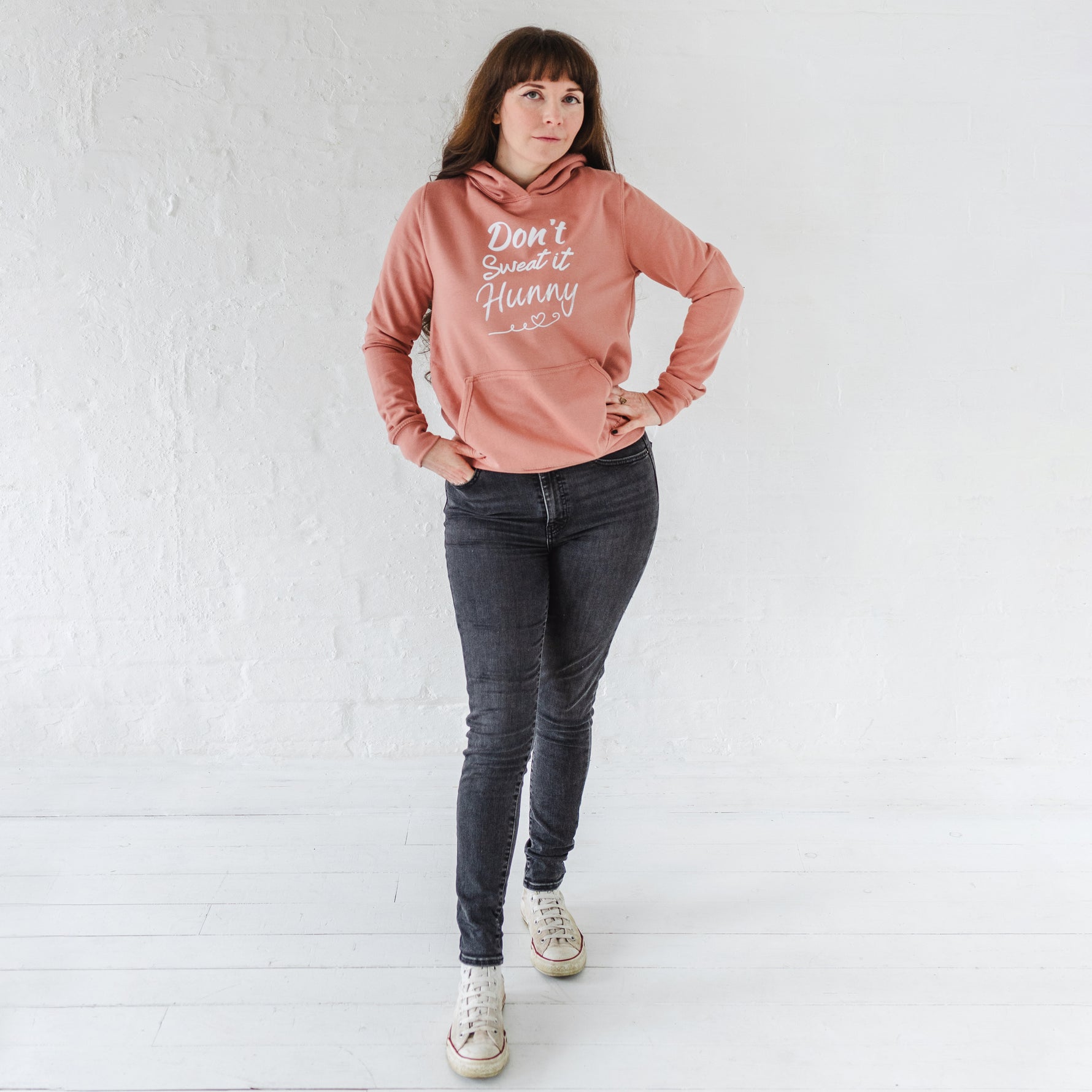 Woman wearing dark grey jeans, sneakers and  dusky pink fitted fashion womens long sleeve hoodie. Hoodie top for workout / casual wear with self empowerment slogan in white writing to front chest reads Don't Sweat It Hunny with line shaped into heart half way and up curl at one end. Ladies fashion hoodie features Hood with fabric drawstrings.