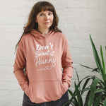 Woman wearing dark grey jeans, sneakers and dusky pink fitted fashion womens long sleeve hoodie. Hoodie top for workout / casual wear with self empowerment slogan in white writing to front chest reads Don't Sweat It Hunny with line shaped into heart half way and up curl at one end. Ladies fashion hoodie features Hands in kangaroo pouch front pocket