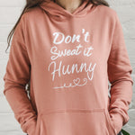 Close up of Woman wearing dusky pink fitted fashion womens long sleeve hoodie. Hoodie top for workout / casual wear with self empowerment slogan in white writing to front chest reads Don't Sweat It Hunny with line shaped into heart half way and up curl at one end. Hands in kangaroo front pocket