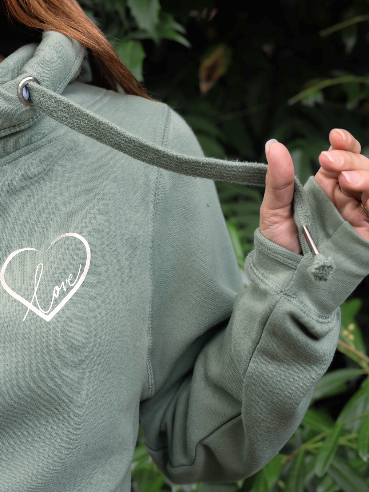 Close up of a woman wearing a ladies hooded sweatshirt with cowl neck, this image shows the cross over neck and cosy thumb hole loops to cuff sleeves - sage green hoodie with thick drawstrings. and a silver metallic love heart design.