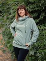 Woman standing by a bush outdoors wearing a Womens Dusty Green Cowl Neck Hoodie - sage green with silver metallic love heart motif, and black leggings. Ladies Hoodie has a cross neck hooded sweatshirt design, long sleeves and thumb hole loops to cuffs.  Thick drawstrings and Front kangaroo pouch pocket. 