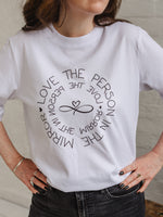 Woman wearing ladies white graphic t-shirt top. Design on fashion Tee reads Love the Person in the Mirror women's relaxed fit white slogan short sleeve boxy oversized t-shirt. Infinity symbol and heart design and circular wording Love the Person in the Mirror in reverse on inner circle and right way round on outer circle so that wording. Mid length sleeve to just above elbows. Worn with black jeans