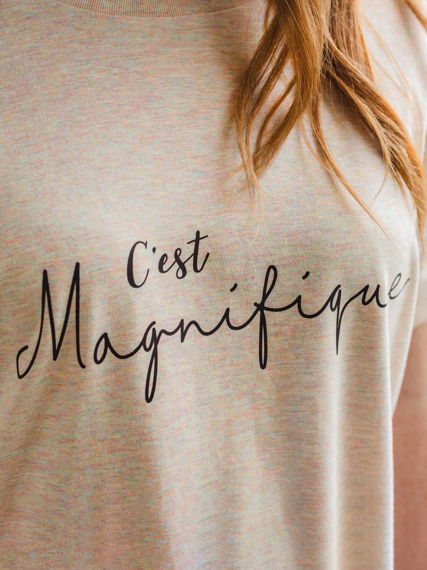 Close up image of womens fashion t shirt in light beige / cream with subtle rainbow multicolour flecks. Black script slogan text across chest reads C'est Magnifique
