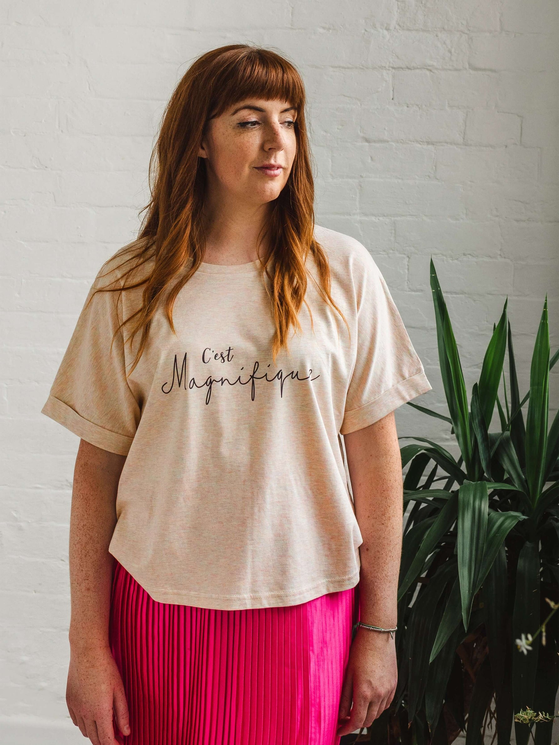 Woman wearing ladies T-Shirt with dolman sleeve, rib neck collar and sleeve cuff fold. Rainbow Heather - light beige/cream base colour with multicolour flecks. Oversized women’s t-shirt with a short sleeves, inspired by the 1990s oversize trend.. Black slogan script font design reads French phrase C'est Magnifique paired with a bright pink satin midi skirt
