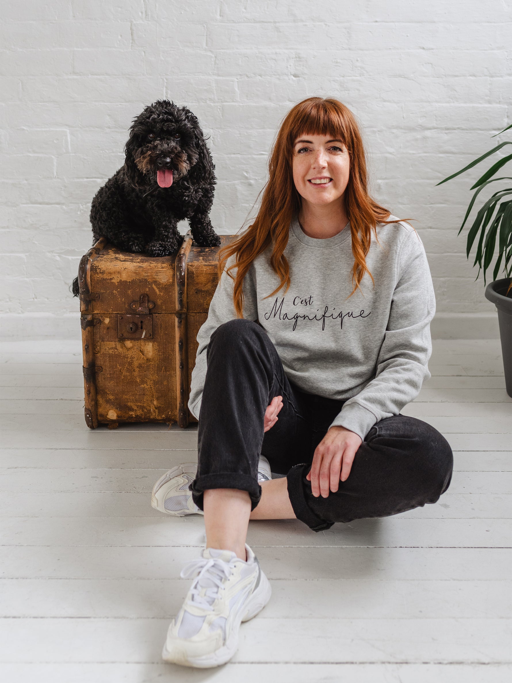 Woman sat on the floor smiling, wearing light grey sweatshirt without hood. Womens jumper unisex relaxed fit with crew neck and long sleeves with cuffs. Model wears black jeans. Slogan in black script font reads French phrase C'est Magnifique, Behind her there is a wooden weathered chest and a black cockapoo dog sitting on it happy and looking to camera with it's tongue out
