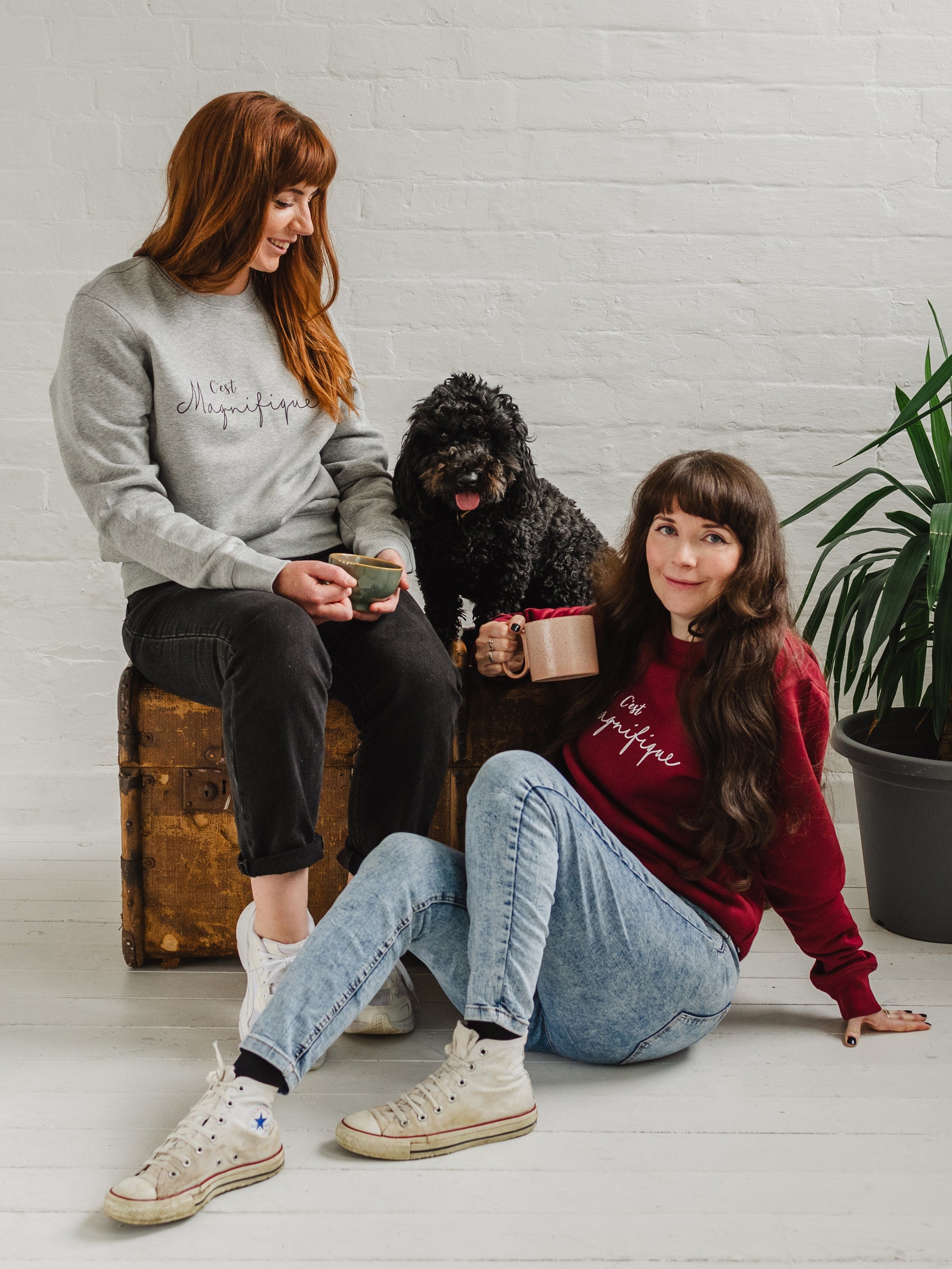 Two women, woman wearing burgundy red sweatshirt is sat on the floor, jumper is unisex relaxed fit with crew neck and long sleeves with cuffs. Model wears light blue acid wash jeans. Slogan in white script font reads French phrase C'est Magnifique, Behind her there is a wooden weathered chest and a black cockapoo dog sitting on it looking forward with it's tongue out. A woman wearing a matching design light grey ladies sweatshirt sits on the chest looking down at the dog smiling. Both hold cups of coffee.