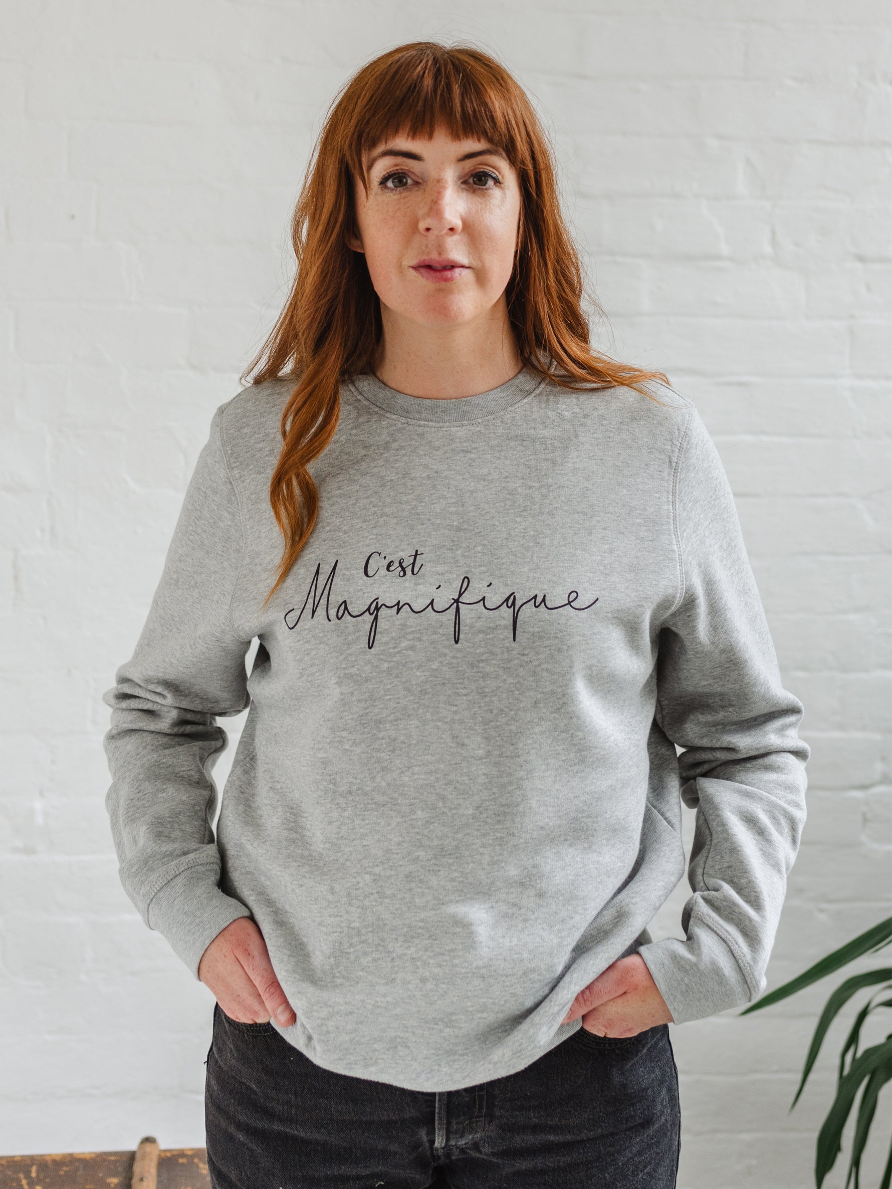 Woman wearing light grey sweatshirt without hood. Womens jumper unisex relaxed fit with crew neck and long sleeves with cuffs. Model wears black jeans. Slogan in black script font reads French phrase C'est Magnifique 