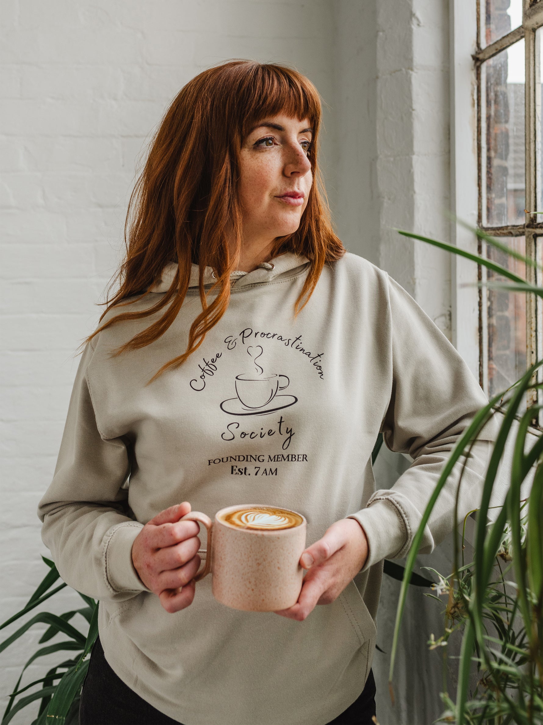 Woman with auburn long hair standing looking out a window holding a mug of coffee wears a Ladies coffee themed oversized fit hoodie. Stone grey hoodie top with coffee cup and saucer design. Black outline with steam from cup shaped into a heart. Writing above reads Coffee & Procrastination. Below reads Society FOUNDING MEMBER Est. 7am.