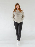 Woman with auburn long hair wears a womens coffee themed oversized fit hoodie. Stone grey hoodie top with coffee cup and saucer design. Black outline with steam from cup shaped into a heart. Writing above reads Coffee & Procrastination. Below reads Society FOUNDING MEMBER Est. 7am. Worn with black straight leg jeans and white trainers 