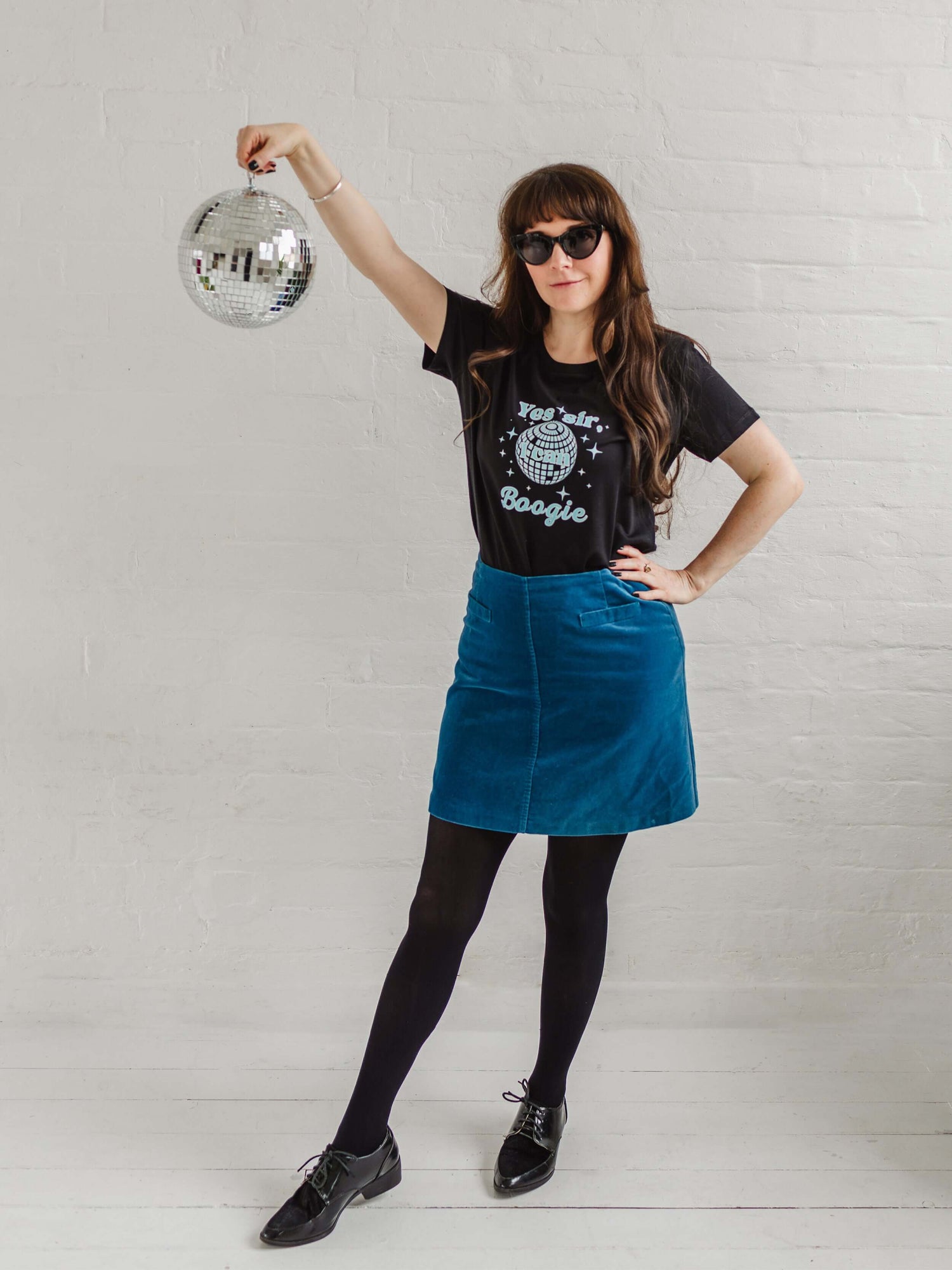 Black graphic t-shirt with mirror ball design.  Woman wearing sunglass smiling holding a silver disco ball, worn with a velvet A line mid blue skirt, black tights and black brogues. Fitted short sleeve tshirt with  round neck. 70s disco music inspired ladies slogan tshirt with disco ball design to background of top and sparkles surround. Slogan text in foreground reads Yes sir I can Boogie in light blue font. Organic cotton casual fitted Tee