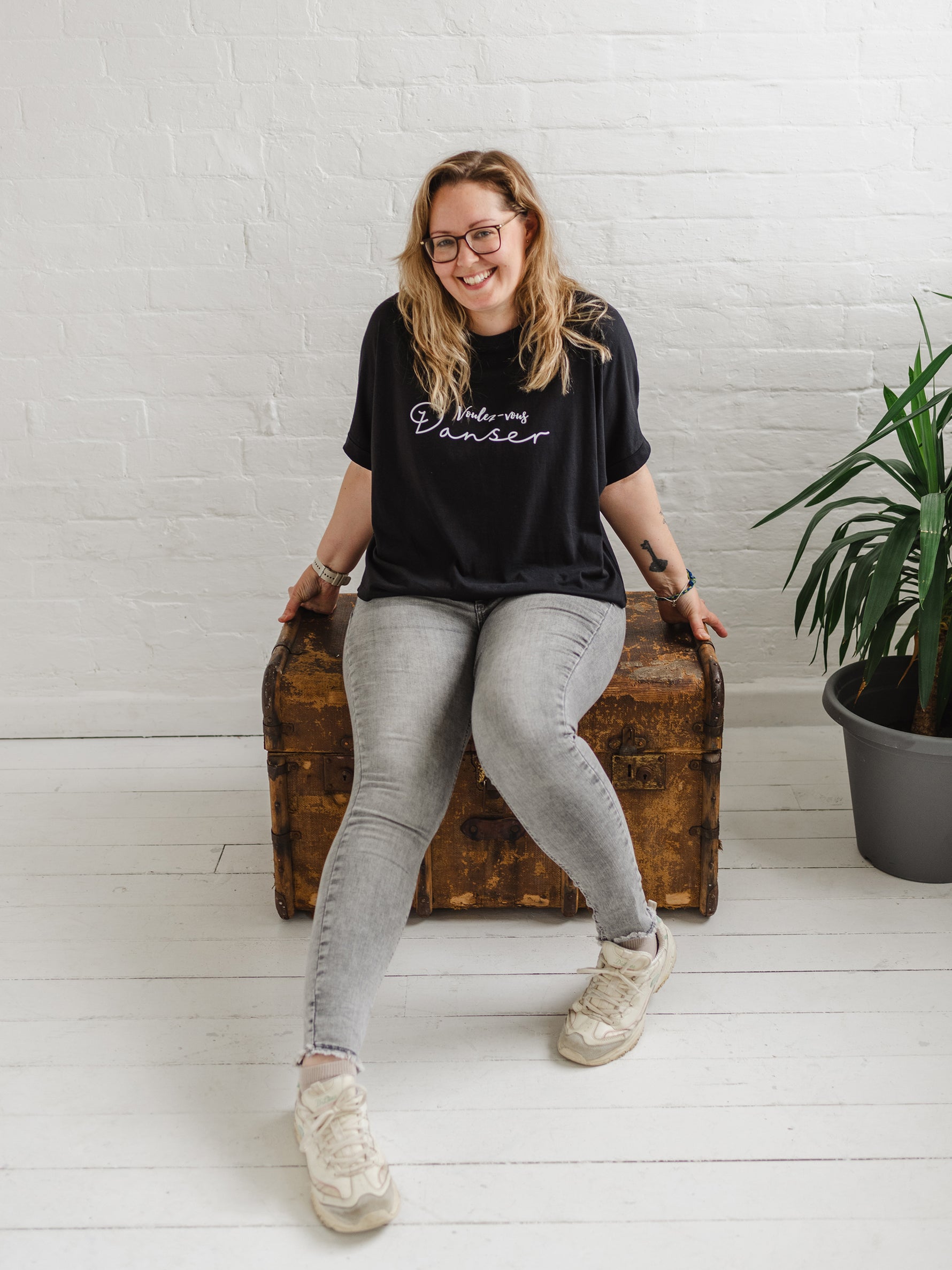Woman wearing a black T-shirt with loose sleeves with roll cuffs worn with grey skinny jeans. The ladies tshirt has the words Voulez Vous Danser in white  She is sitting on a wooden weathered chest and is wearing white trainers