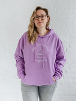 Woman with blonde long hair standing wears a womens coffee themed oversized fit hoodie. Lavender purple hoodie top with coffee cup and saucer design. White outline with steam from cup shaped into a heart. Writing above reads Coffee & Procrastination. Below reads Society FOUNDING MEMBER Est. 7am. She pairs it with grey skinny jeans