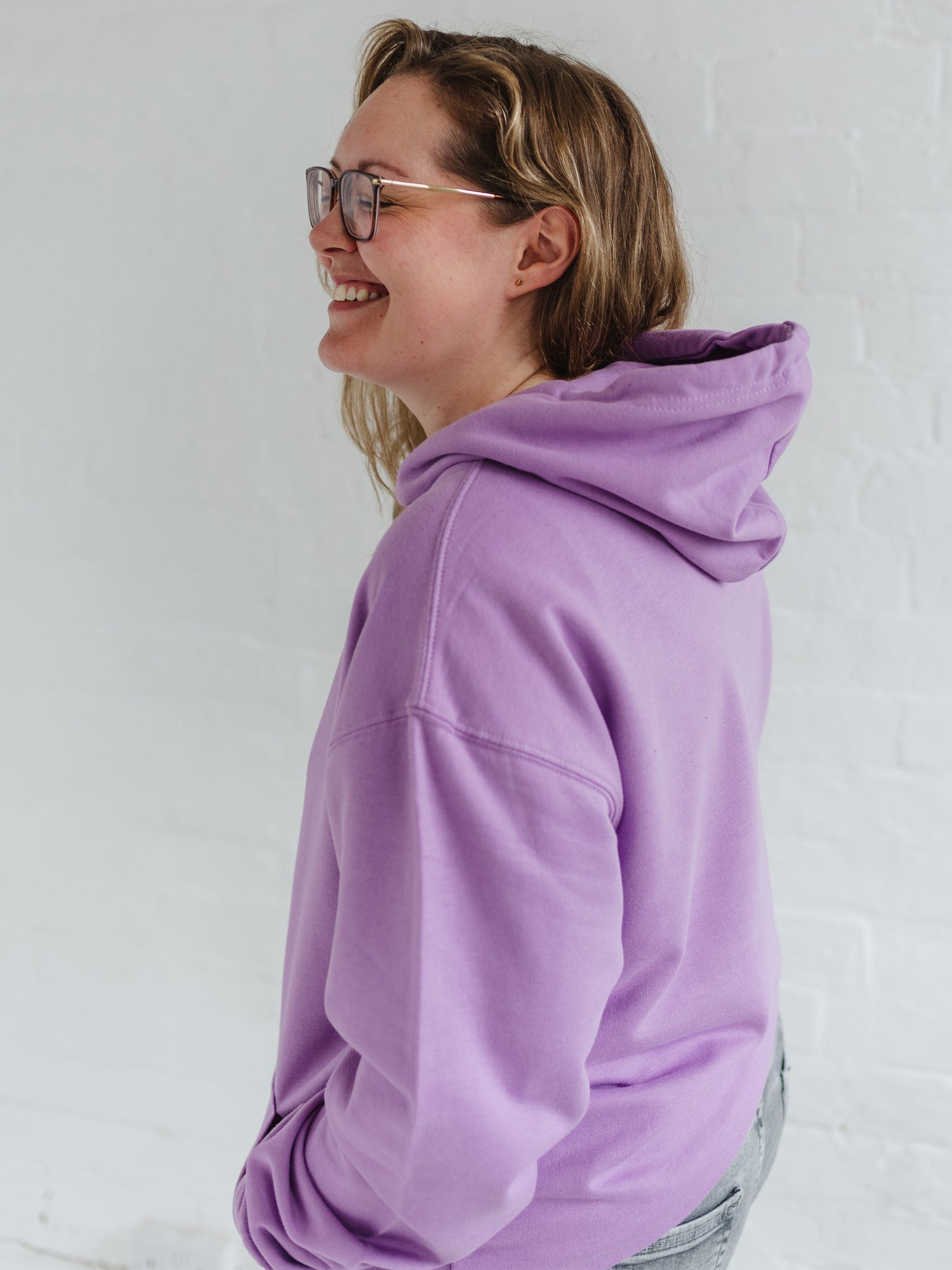 Woman profile smiling has blonde long hair and glasses  wears a Ladies coffee themed relaxed unisex fit hoodie. Lavender purple long sleeve pullover hoodie top with coffee cup and saucer design. White outline with steam from cup shaped into a heart. Writing above reads Coffee & Procrastination. Below reads Society FOUNDING MEMBER Est. 7am. She pairs it with grey skinny jeans 