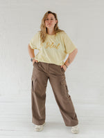 Woman with blonde hair and glasses, wearing butter light yellow oversized t-shirt with loose dolman sleeves with rolled tacked cuff and round neckline loosely tucked into taupe cargo trousers.. Ladies t shirt has the word design Le Soleil across the chest in matt metallic gold script font.