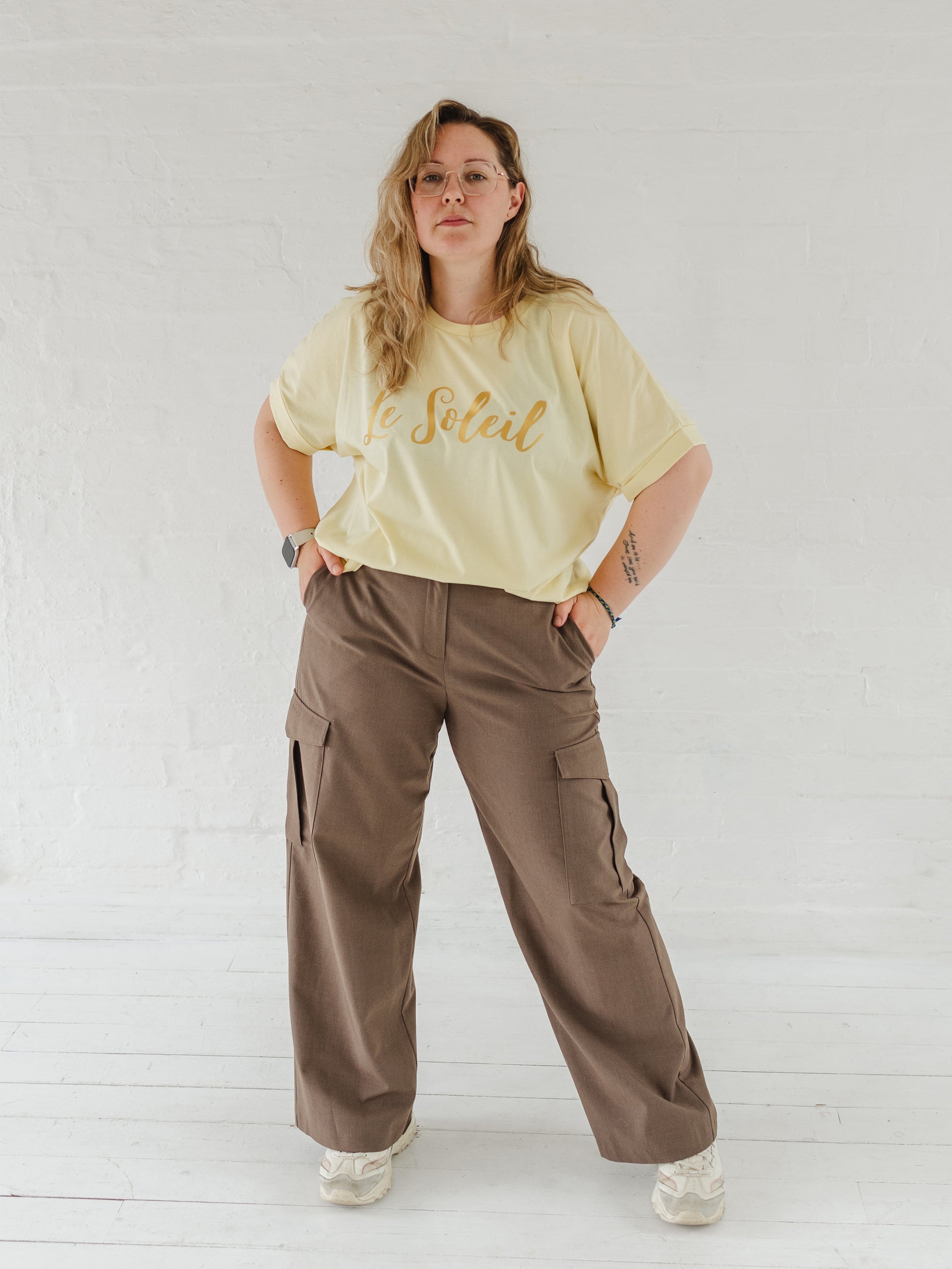 Woman with blonde hair and glasses, wearing butter light yellow oversized t-shirt with loose dolman sleeves with rolled tacked cuff and round neckline loosely tucked into taupe cargo trousers.. Ladies t shirt has the word design Le Soleil across the chest in matt metallic gold script font.