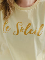 Close up of woman with blonde hair wearing butter light yellow oversized t-shirt with loose dolman sleeves with rolled tacked cuff and round neckline. Ladies t shirt has the word design Le Soleil across the chest in matt metallic gold script font.