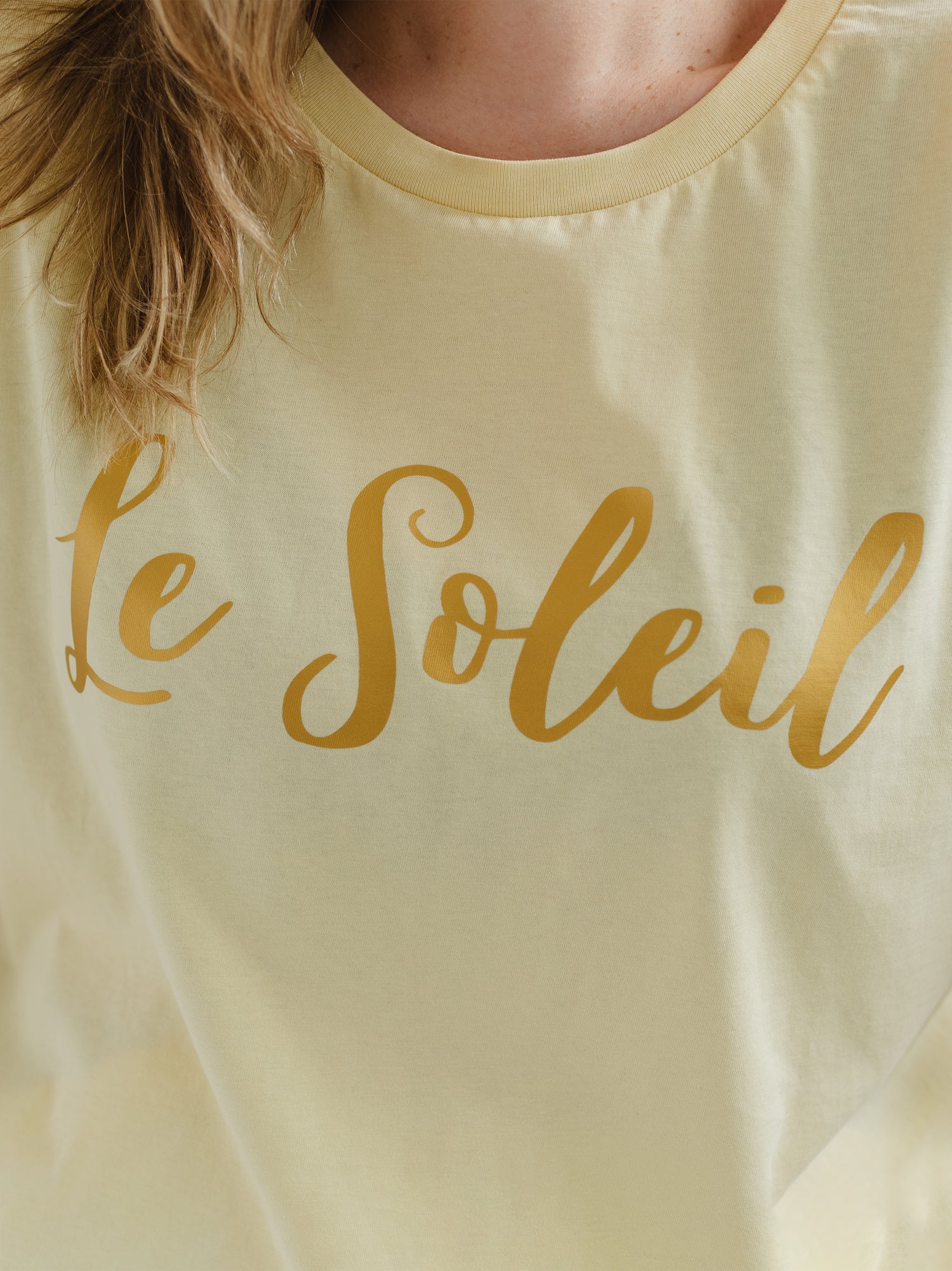Close up of woman with blonde hair wearing butter light yellow oversized t-shirt with loose dolman sleeves with rolled tacked cuff and round neckline. Ladies t shirt has the word design Le Soleil across the chest in matt metallic gold script font.