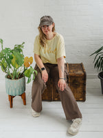 Woman sitting on a wooden weathered chest. Blonde hair and glasses, wearing butter light yellow oversized t-shirt with loose dolman sleeves with rolled tacked cuff and round neckline loosely tucked into taupe cargo trousers.. Ladies t shirt has the word design Le Soleil across the chest in matt metallic gold script font. Woman wears taupe casual baseball cap
