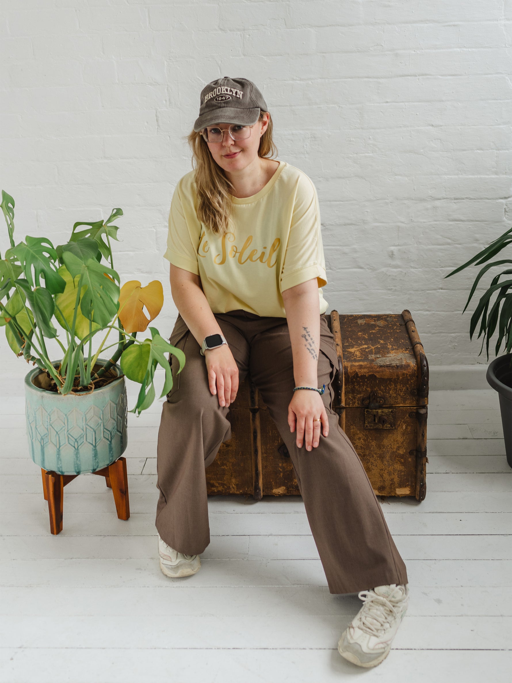 Woman sitting on a wooden weathered chest. Blonde hair and glasses, wearing butter light yellow oversized t-shirt with loose dolman sleeves with rolled tacked cuff and round neckline loosely tucked into taupe cargo trousers.. Ladies t shirt has the word design Le Soleil across the chest in matt metallic gold script font. Woman wears taupe casual baseball cap