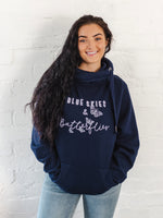 Ladies Navy blue hoodie with cross cowl neck. Thumb holes to sleeves. Design is of four light grey butteflies in flight and wording Blue Skies and Butterflies to the front. Unisex relaxed fit hoodie. Light purple wording with light grey back shadow. Long sleeves and straps from cowl neck with metal toggles on the end. Worn by caucasian woman with long dark brown hair smiling . Worn with blue jeans