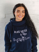 Womens Navy blue hoodie with cross cowl neck. Thumb holes to sleeves. Design is of four light grey butteflies in flight and wording Blue Skies and Butterflies to the front. Unisex relaxed fit hoodie. Light purple wording with light grey back shadow. Long sleeves and straps from cowl neck with metal toggles on the end. Worn by caucasian woman with long dark brown hair smiling 