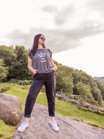 Woman wearing a grey graphic t-shirt and dark jeans standing on a rock with a scenic background. Summer Festival Women's Band Tee. Ladies T shirt Grey cotton cap sleeve t shirt with lightning bolt design and slogan I'm with the Band. 