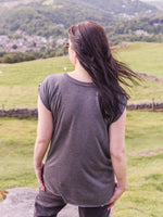 Woman outside wearing sunglasses. Woman outside in field. Summer Festival Women's Band Tee. Ladies T shirt Grey cotton cap sleeve t shirt . Back view shows lower hem design at the back sits below bottom. Round crew neck 