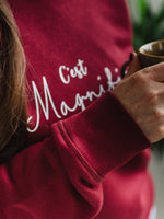 Close up of womens burgundy red trendy sweatshirt with white slogan script wording C'est Magnifique. Woman is holding a coffee cup 