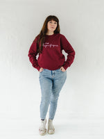 Full length photo of a caucasian woman in her 40s with long brown hair wearing light blue acid wash jeans and a burgundy red on trend ladies sweatshirt, oversized fit with crew neck and long sleeves.