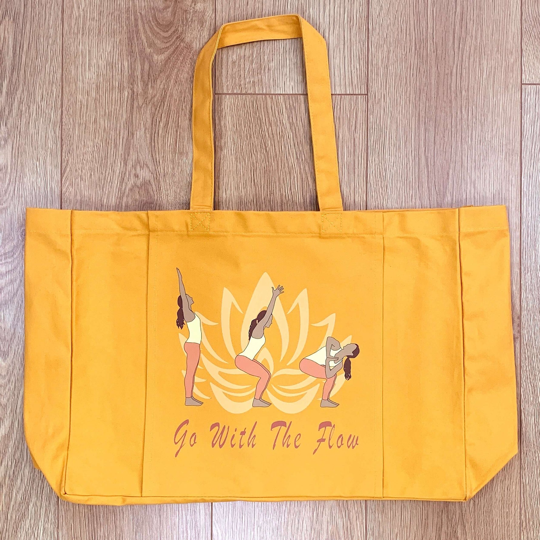 An amber coloured yoga bag on the wooden floor of a fitness studio. The bag hosts the words Go With The Flow and a graphic of a lotus flower and 3 yoga poses.