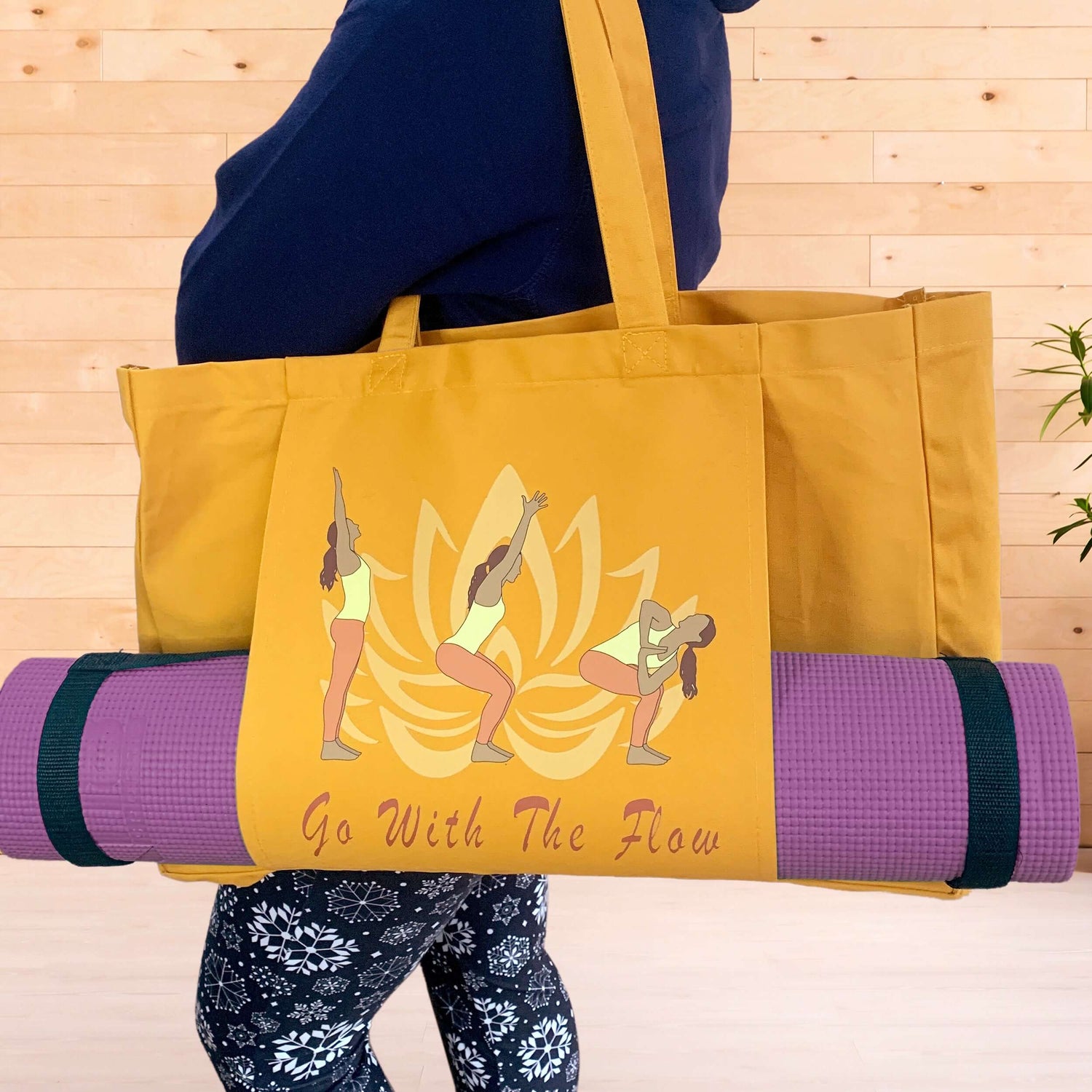 A woman in workout wear hoodie and leggings holding an amber coloured yoga bag on her shoulder in a fitness studio. The yoga mat bag hosts the words Go With The Flow and a graphic of a lotus flower and a dark skinned woman in 3 yoga poses. A purple yoga mat is held by a pouch pocket on the front of the bag.