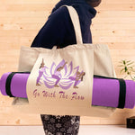 A woman holding a natural coloured yoga bag on her shoulder in a fitness studio. The yoga mat bag hosts the words Go With The Flow and a graphic of a lotus flower and dark skinned long haired woman in 3 yoga poses. A purple yoga workout mat is held by a pouch pocket on the front of the bag.Large organic cotton vegan approved shoulder bag.