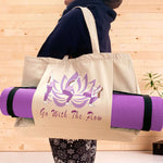 A relaxed woman holding a natural coloured yoga bag on her shoulder in a fitness studio. The bag hosts the words Go With The Flow and a graphic of a lotus flower and 3 yoga poses. A purple yoga mat is held by a pouch pocket on the front of the bag.