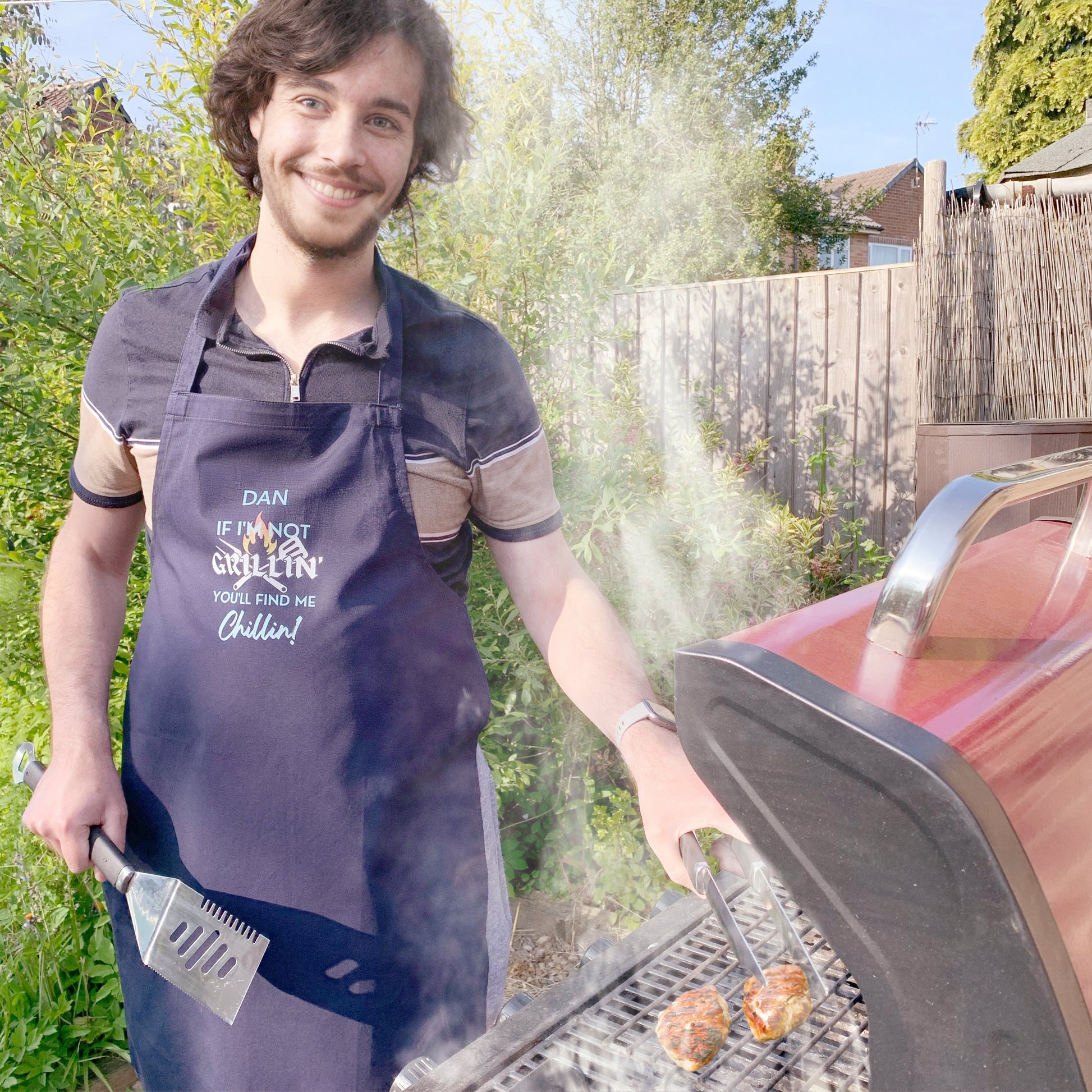 Man stood in garden at a barbecue cooking wearing dark navy blue cooking apron. Personalised with name and slogan writing If I'm not Grillin' you'll find me Chillin! BBQ grill cooking utensil behind text design. 