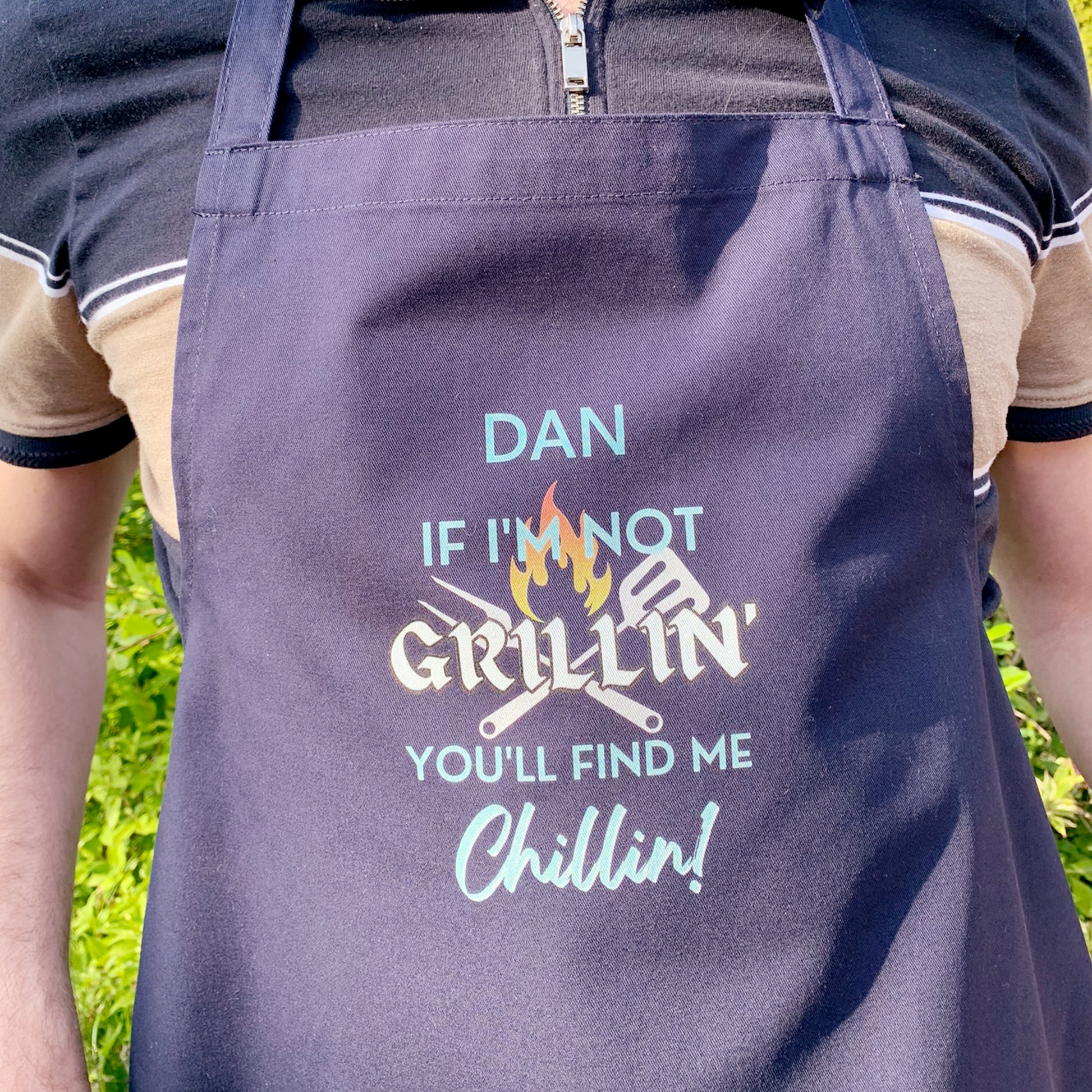 Man wearing dark navy blue cooking apron. Personalised with name and slogan writing If I'm not Grillin' you'll find me Chillin!  BBQ cooking utensil behind text design.    A Perfect gift for him for Father's day of birthday 