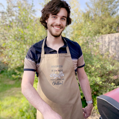 Man stood at barbecue wearing khaki navy blue cooking apron. Personalised with name and slogan writing If I'm not Grillin' you'll find me Chillin! BBQ cooking utensil behind text design