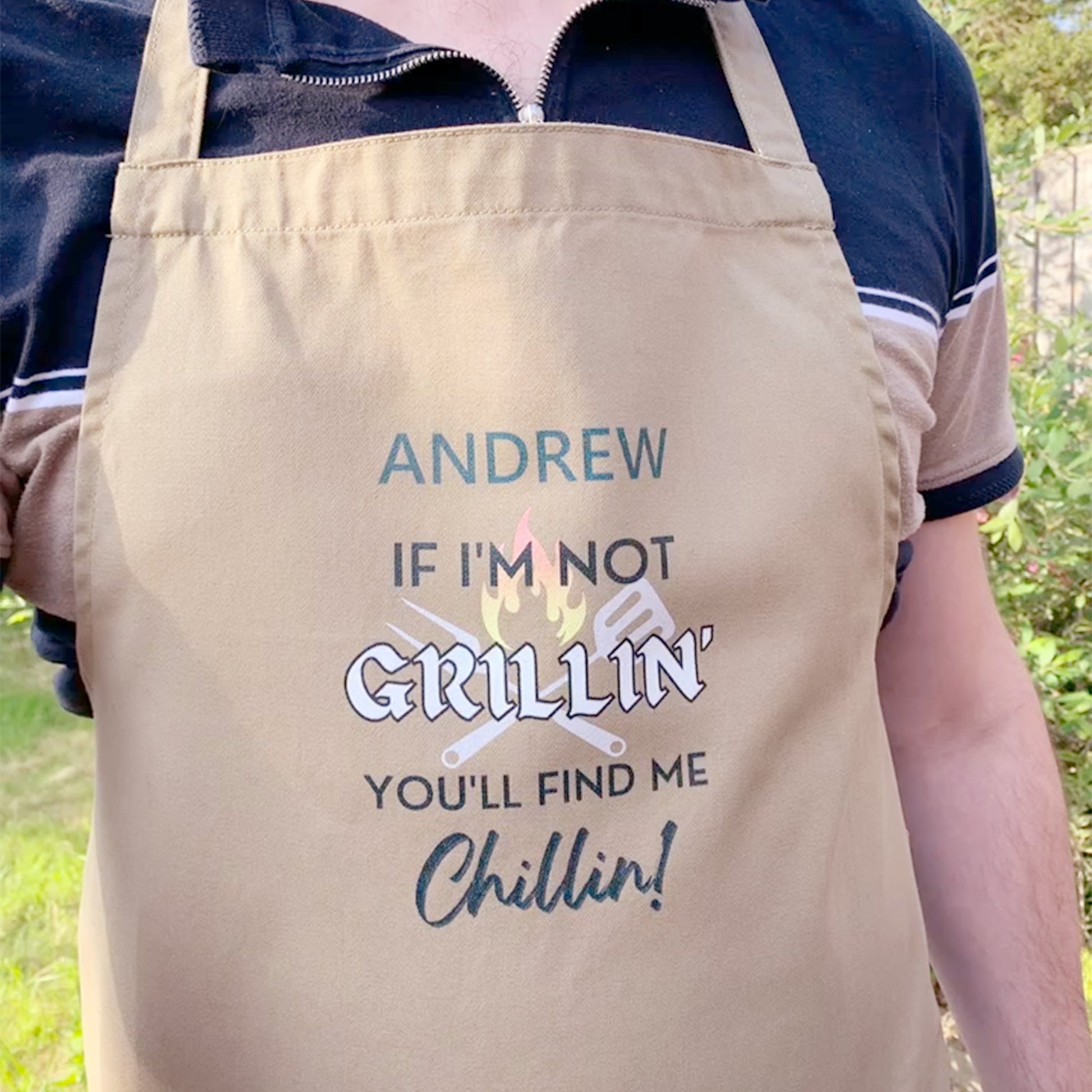 Man wearing khaki cooking apron. Personalised with name and slogan writing If I'm not Grillin' you'll find me Chillin! BBQ cooking utensil and flame behind text design. 