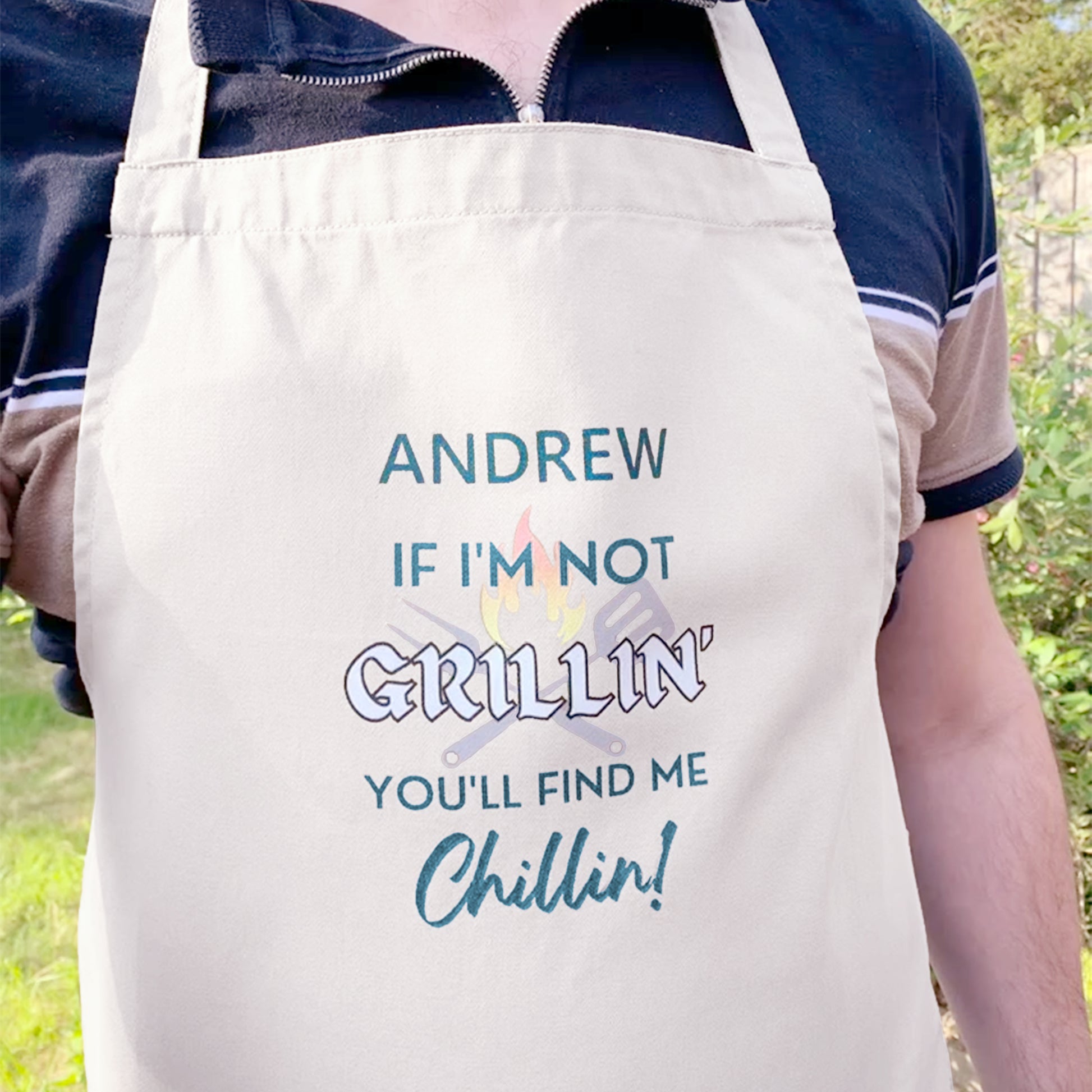 Man wearing Off white cooking apron. Personalised with name and slogan writing If I'm not Grillin' you'll find me Chillin! BBQ cooking utensil and flame behind text design. 