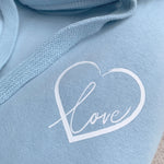 Close up photo of sky blue unisex Love heart cross cowl neck casual fashion hoodie. Cotton Polyster blend relaxed fit unisex oversized hoodie design has love heart. White heart to upper left of the chest with word Love in white handwriting font nestled within the heart.