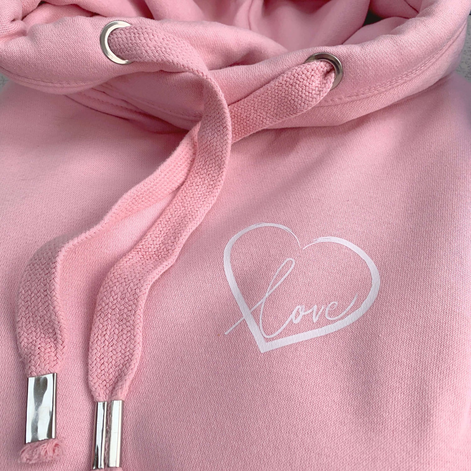 Close up to baby pink womens hoodie Love heart cross cowl neck casual fashion hoodie. Cotton Polyester blend relaxed fit unisex oversized style hoodie design has love heart. Whitert to upper left of the chest with word Love in white script font nestled within the heart.