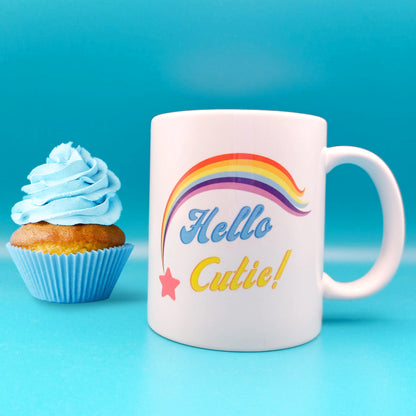 White tea / coffee mug with cute pink shooting star and rainbow trail about the words Hello Cutie blue and yellow slogan wording. Blue background with cupcake to left of the coffee cup
