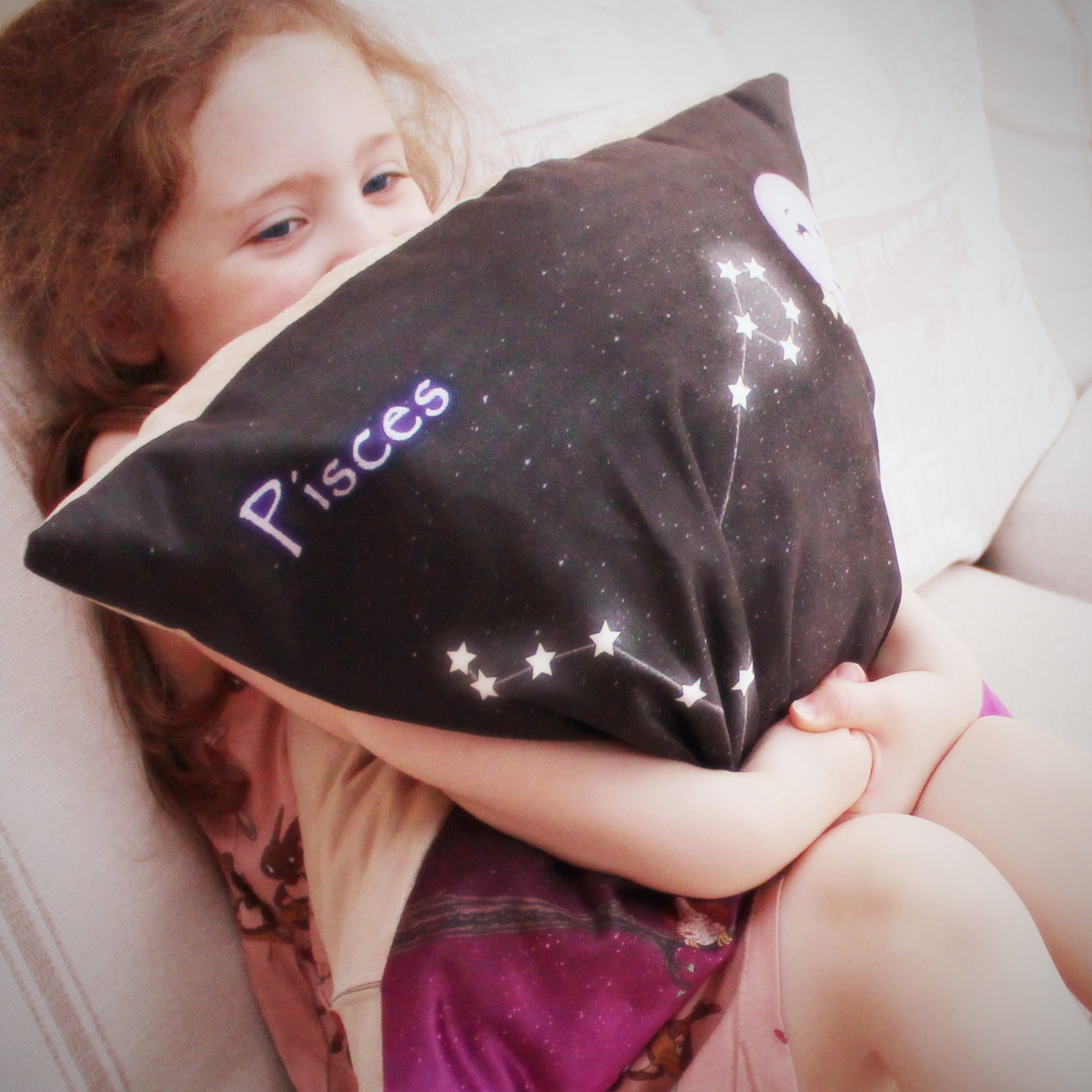 Litte girl hugging Personalised cushion cover with zodiac sign Pisces. Space backdrop black sky with stars fading to deep pink purple hue at bottom of the cushion. Personalised with name at the bottom, Small owl sitting on a branch to the left of star sign constellation at centre and top right cute smiling moon and two stars. 18 inch faux suede home decor constellation cushion. Light Beige sude effect backing to cover