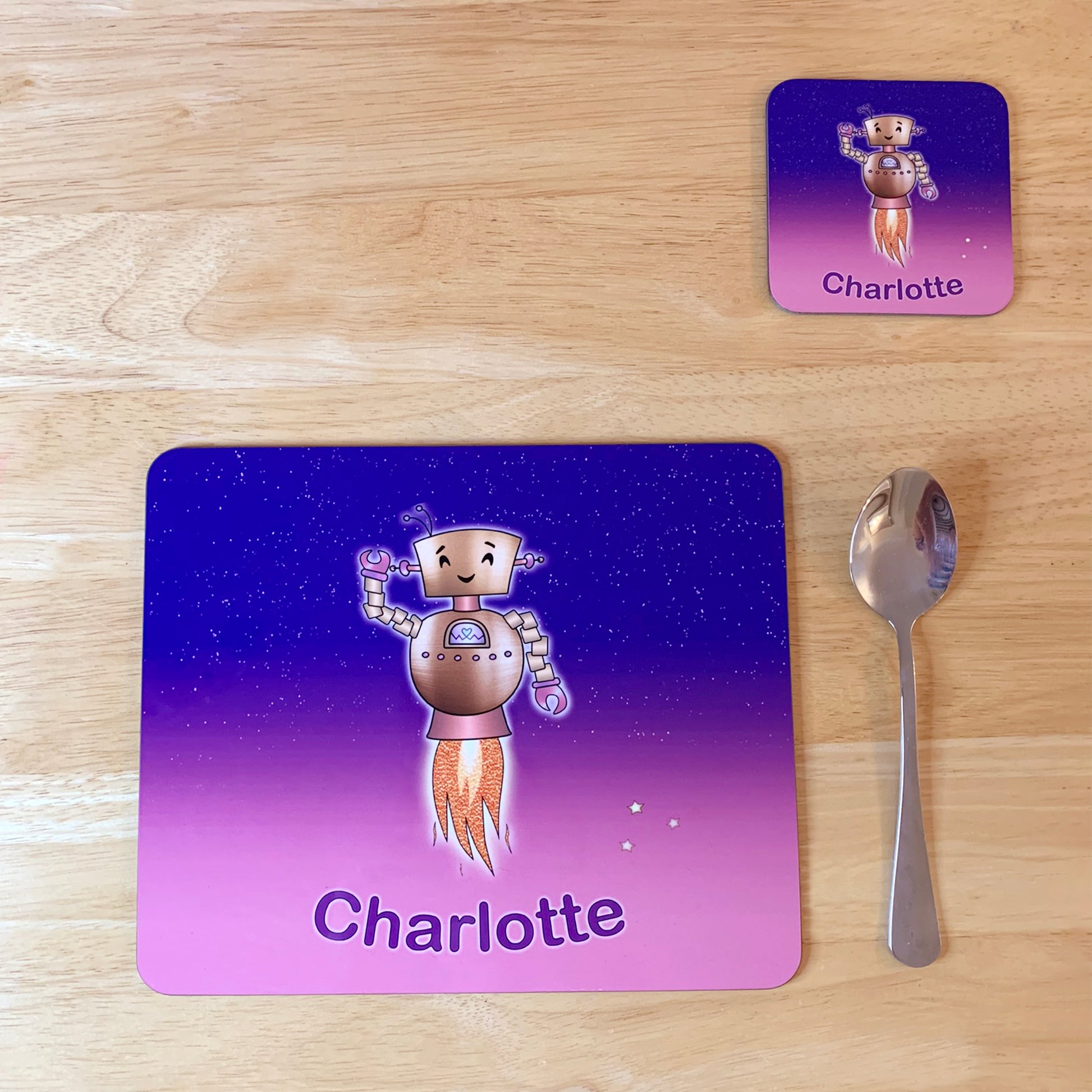 Pink and purple star sky kids placemat set. Robot smiling with jets flying into space. Personalised with girl's name at the bottom of custom child's dinner mat and matching coaster.  Kids spoon next to mat